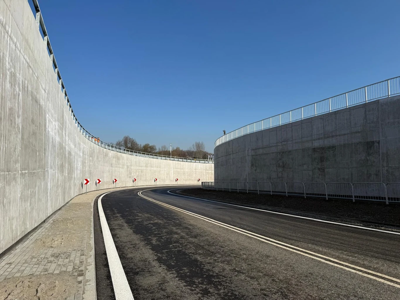 nowoczesna droga o gładkiej nawierzchni otoczona wysokimi betonowymi ścianami, zakręcająca w lewo z widocznymi znakami ostrzegającymi o zakręcie na tle bezchmurnego nieba