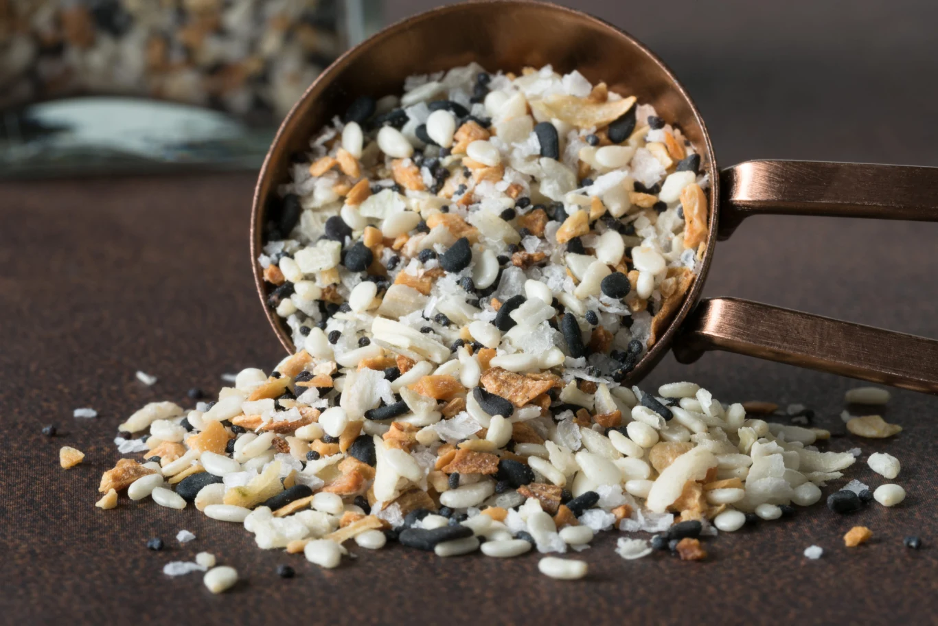 Pyszna i zdrowa przyprawa do pieczywa i nie tylko Mieszanka białego i czarnego sezamu, suszonej cebuli i czosnku wysypana z metalowej miarki na ciemnobrązowym tle.