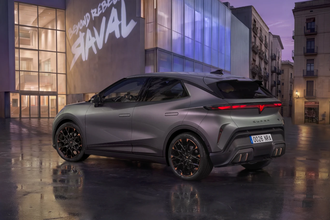 nowoczesny, sportowy SUV o matowym, szarym lakierze i czarnych felgach, podświetlony światłami miejskiej architektury; auto stoi na połyskującej nawierzchni, w tle przeszklony budynek z napisem „BEYOND REBELS STAVAL” oraz zabudowa miejska