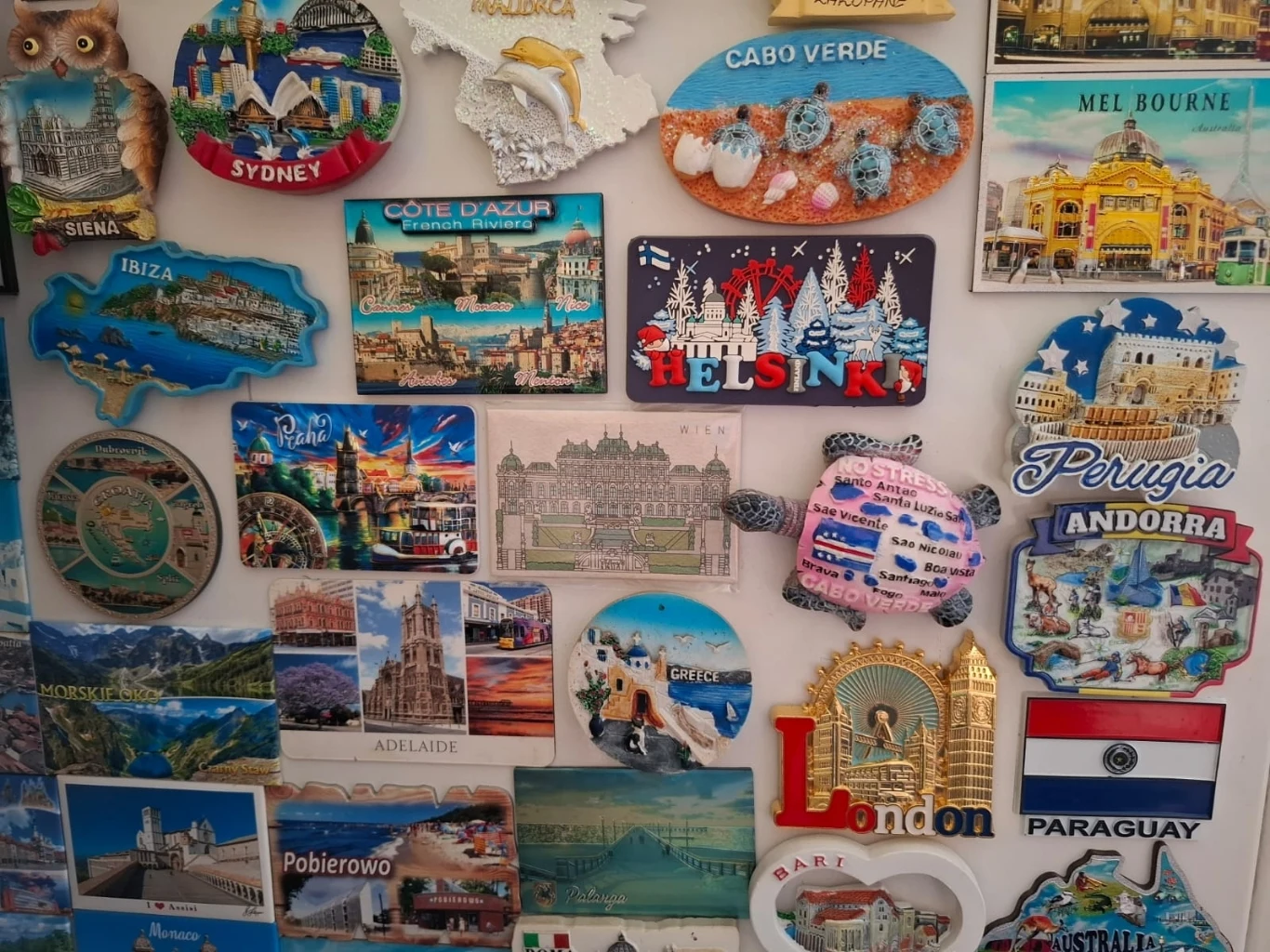 Różnokolorowa kolekcja magnesów z nazwami i wizerunkami miast oraz państw z całego świata, ułożona na białej powierzchni, przedstawiająca charakterystyczne zabytki i krajobrazy każdego miejsca.