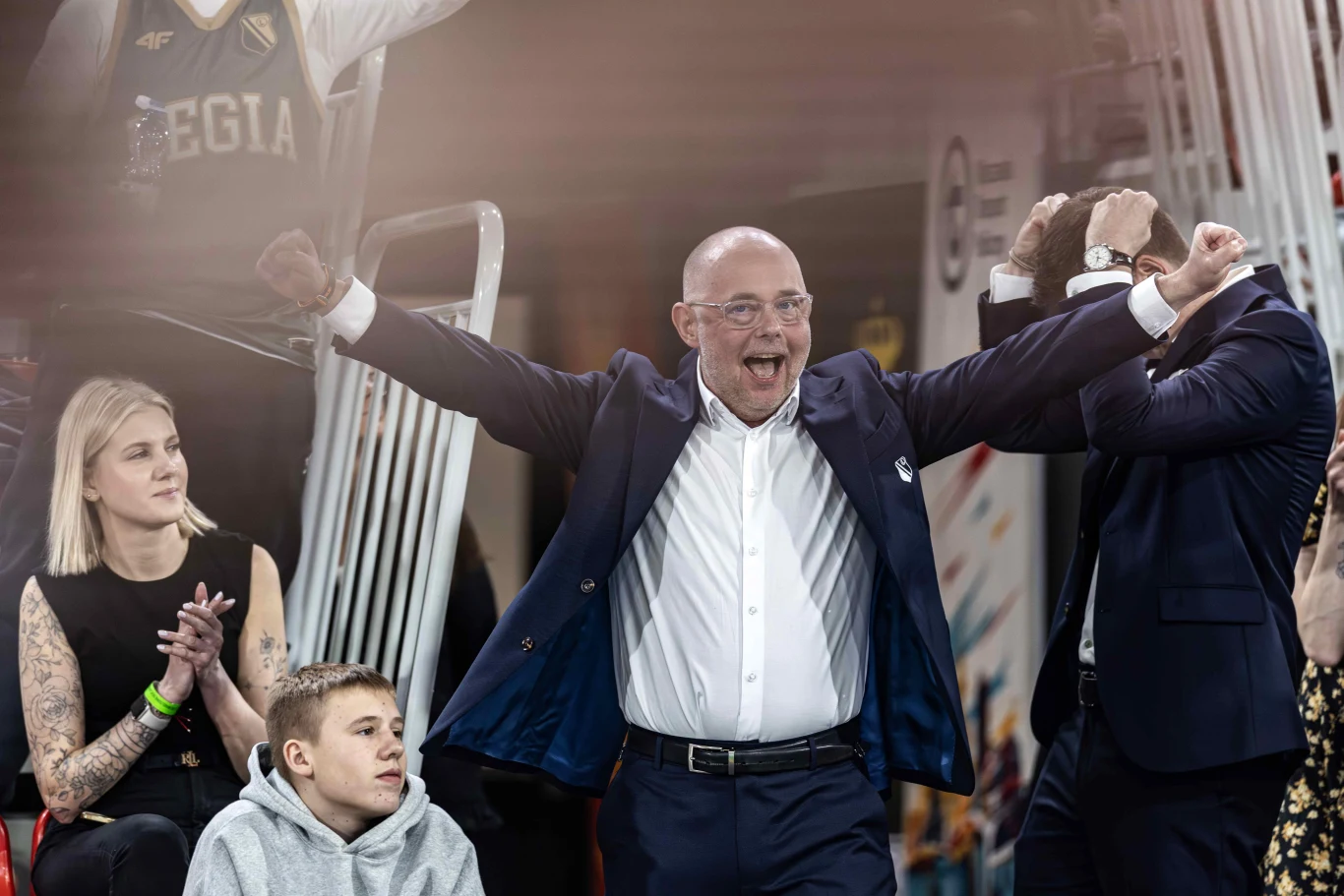 mężczyzna w garniturze z uniesionymi ramionami wyraża radość i entuzjazm, obok niego siedzi kobieta z tatuażami na rękach oraz chłopiec w szarej bluzie, w tle kibice ubrane w stroje z napisem Legia