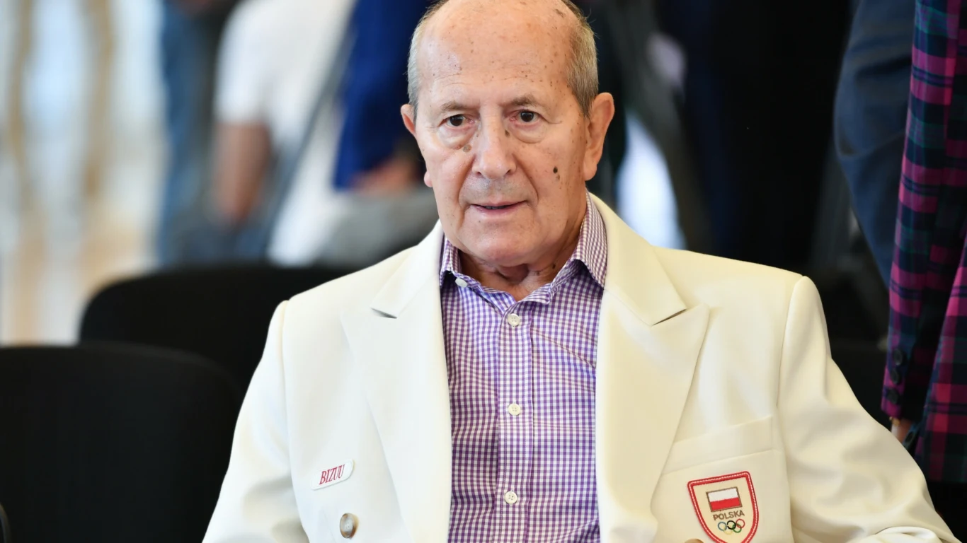 starszy mężczyzna w białej marynarce z emblematem Polskiego Komitetu Olimpijskiego siedzi na czarnym krześle, ma na sobie fioletową koszulę w kratę, patrzy prosto przed siebie