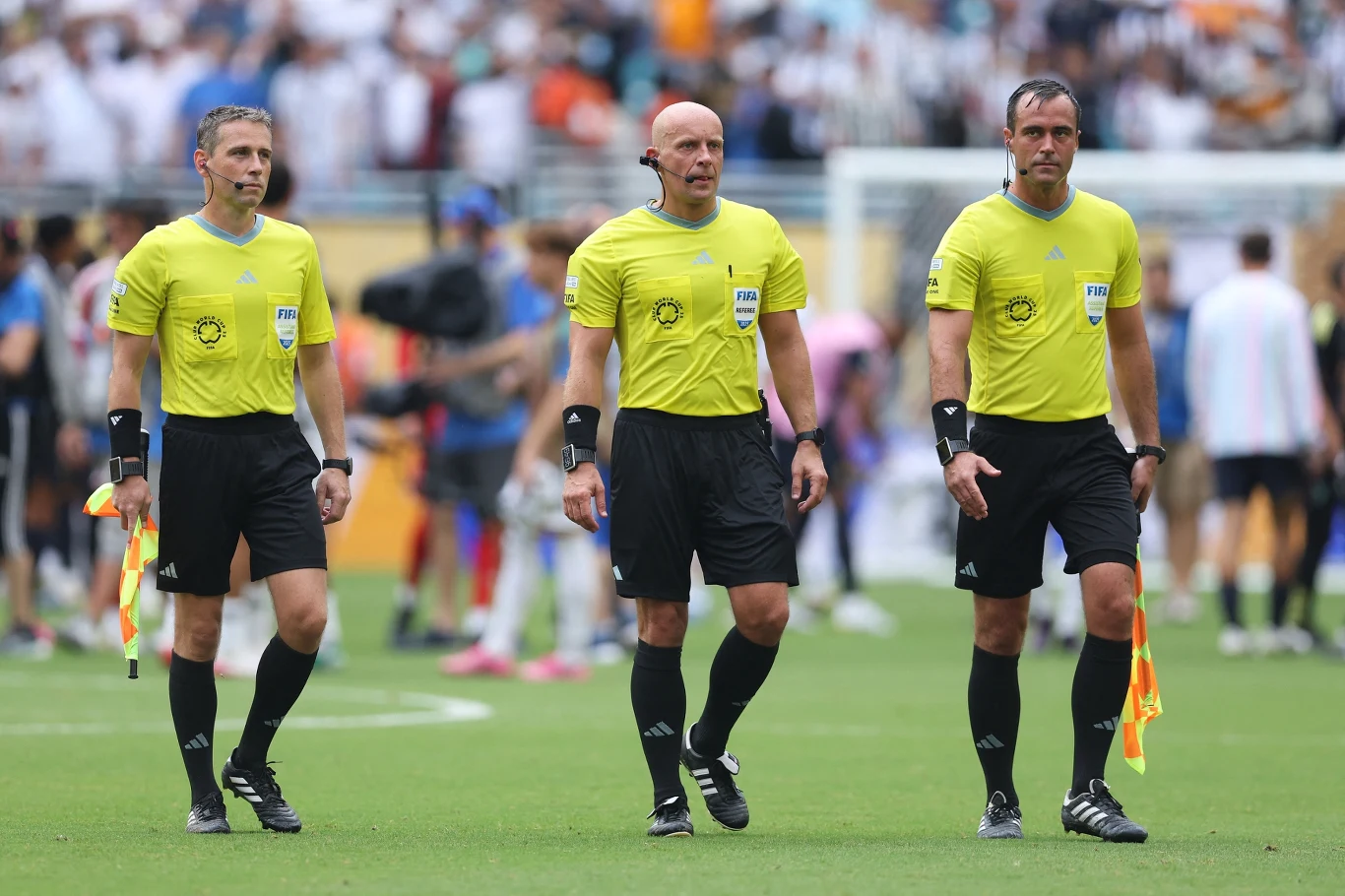 Trzech sędziów piłkarskich w żółtych koszulkach i czarnych spodenkach idzie razem po murawie boiska, dwaj z nich trzymają chorągiewki sędziowskie, w tle rozmazani zawodnicy i kibice.