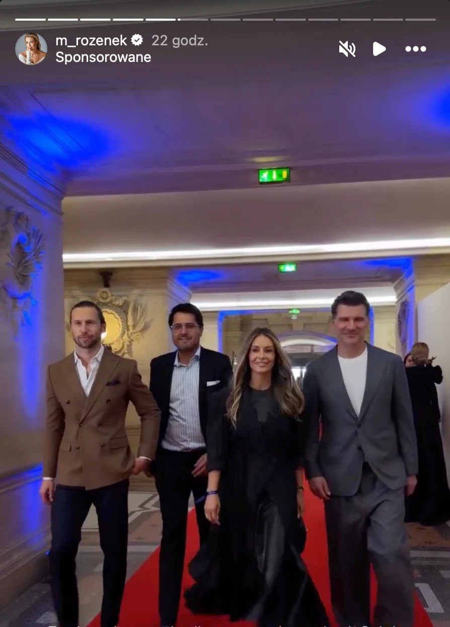 czterech elegancko ubranych dorosłych idzie po czerwonym dywanie w oświetlonym na niebiesko korytarzu o pałacowym wystroju, kobieta w długiej czarnej sukni w towarzystwie trzech mężczyzn w garniturach