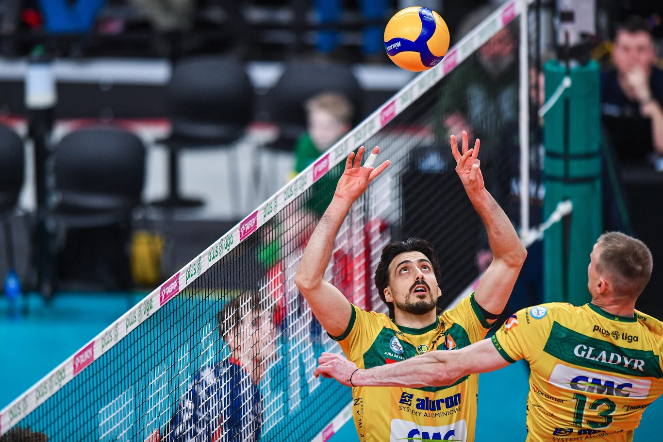 dwóch siatkarzy drużyny w żółto-zielonych strojach tuż przy siatce, jeden zawodnik przygotowuje się do rozegrania piłki do ataku, drugi oczekuje na piłkę, a nad nimi unosi się piłka do siatkówki
