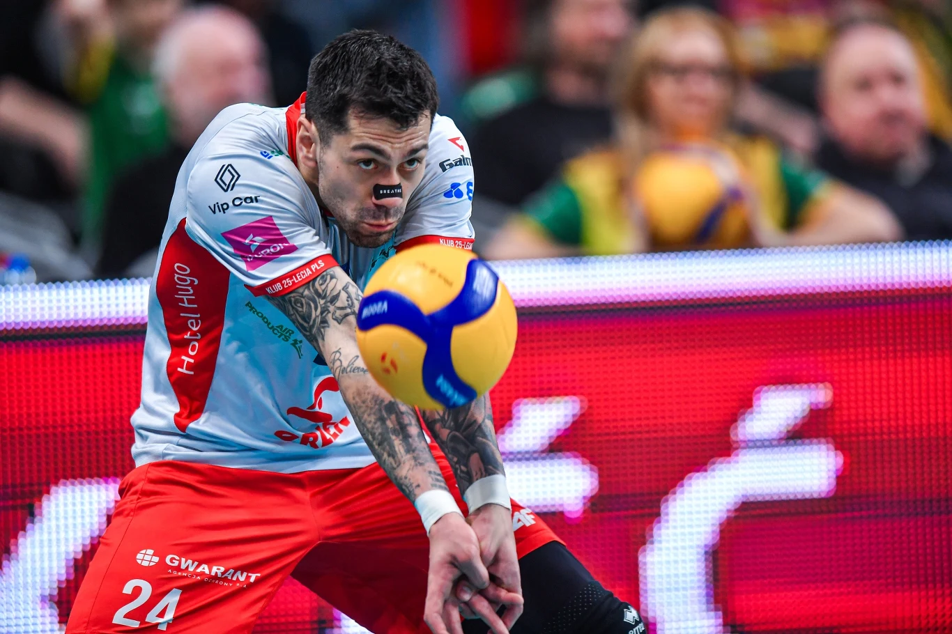 siatkarz w czerwono-białym stroju z numerem 24 odbijający piłkę podczas meczu, skupiona twarz i dynamiczna postawa podkreślają intensywność sportowej rywalizacji