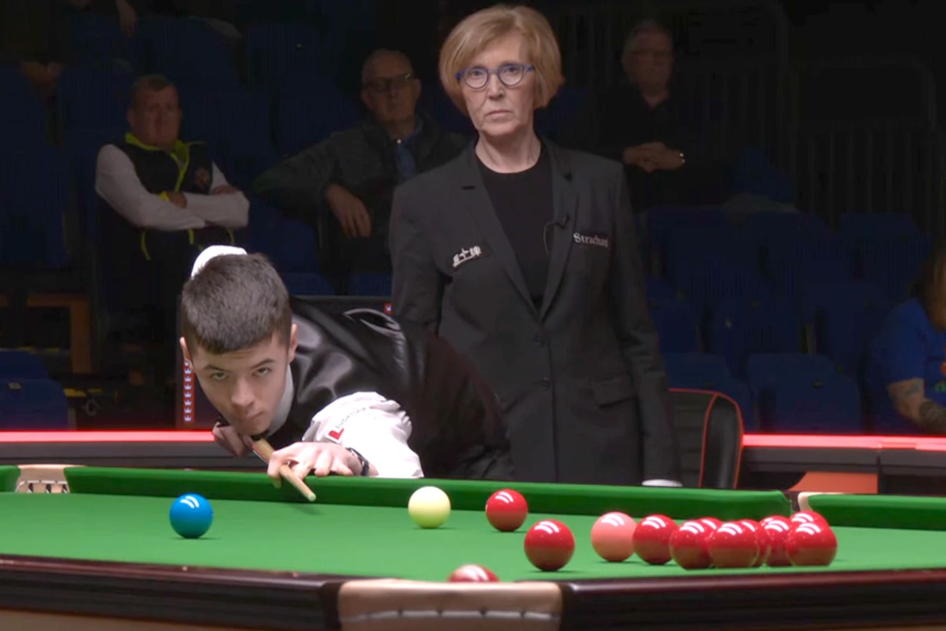 zawodnik skupiony nad uderzeniem w bilardzie snookerowym, przy stole znajduje się sędzia uważnie obserwująca przebieg gry, wokół widoczni są nieliczni widzowie na trybunach