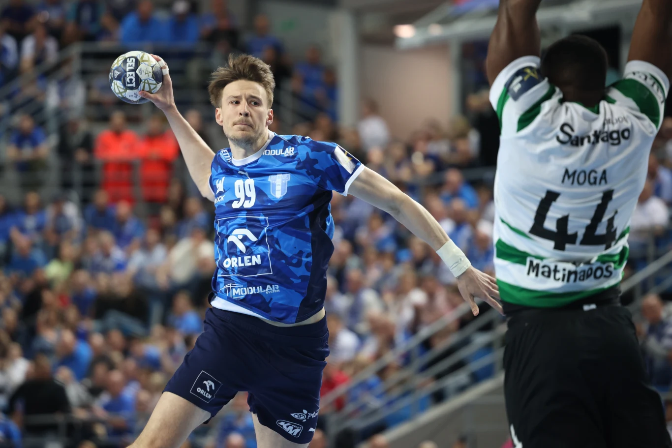 zawodnik w niebieskiej koszulce z numerem 99 wykonuje rzut podczas dynamicznej akcji w meczu piłki ręcznej, obok niego obrońca w biało-zielonej koszulce z wyciągniętymi rękami próbuje zablokować piłkę, wokół widać skupioną publiczność w tle