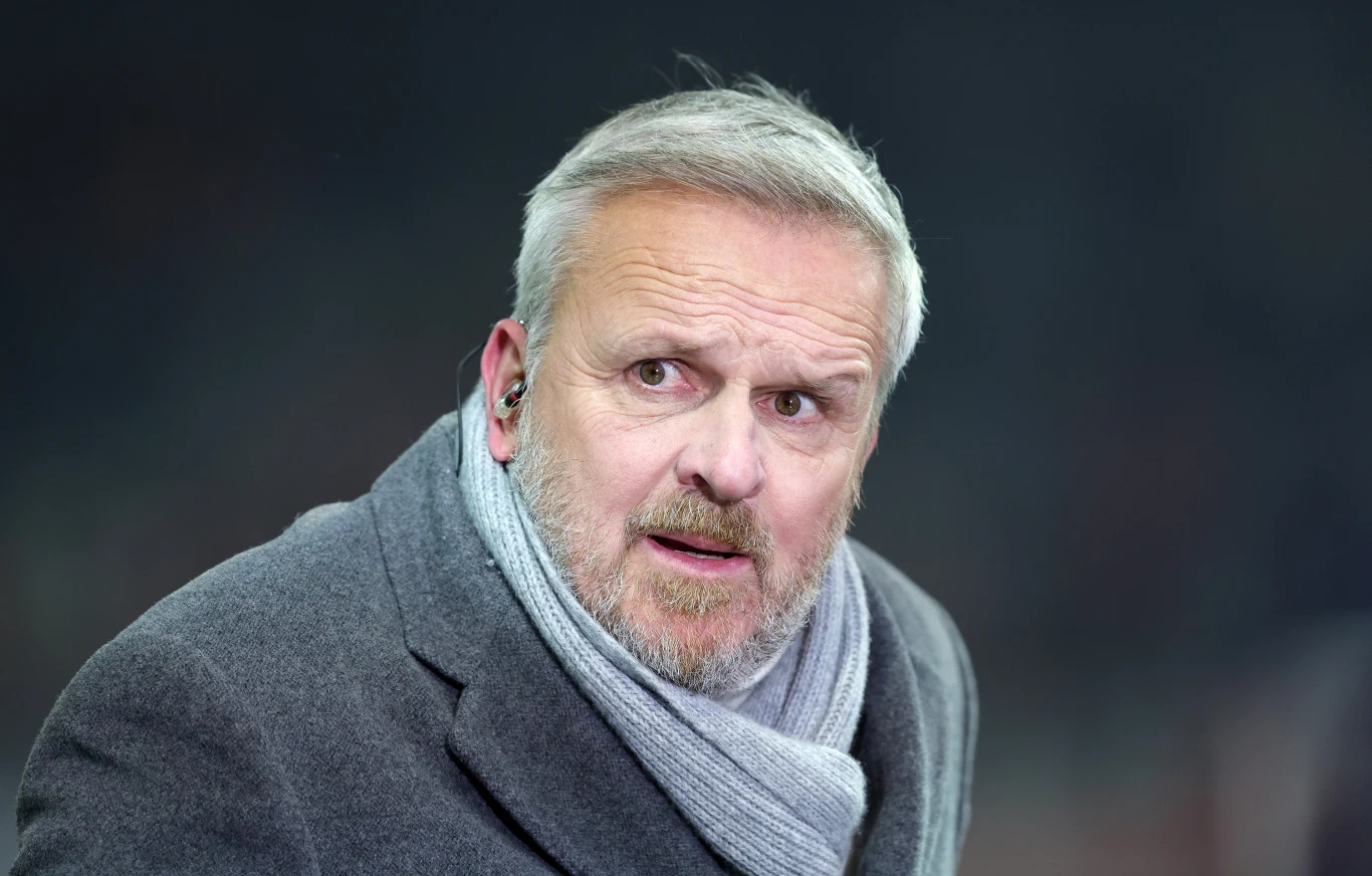 mężczyzna w średnim wieku z siwą brodą i włosami, ubrany w szary płaszcz oraz szalik, z założoną słuchawką w uchu na tle rozmytego, ciemnego tła