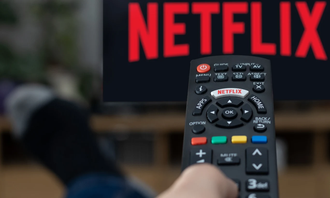 Czy Netflix będzie musiał wypłacić użytkownikom poszczególnych planów zwrot kosztów za podwyżki? Pilot skierowany w stronę ekranu telewizora z logo Netflix, w tle rozmyta postać siedząca przed TV.