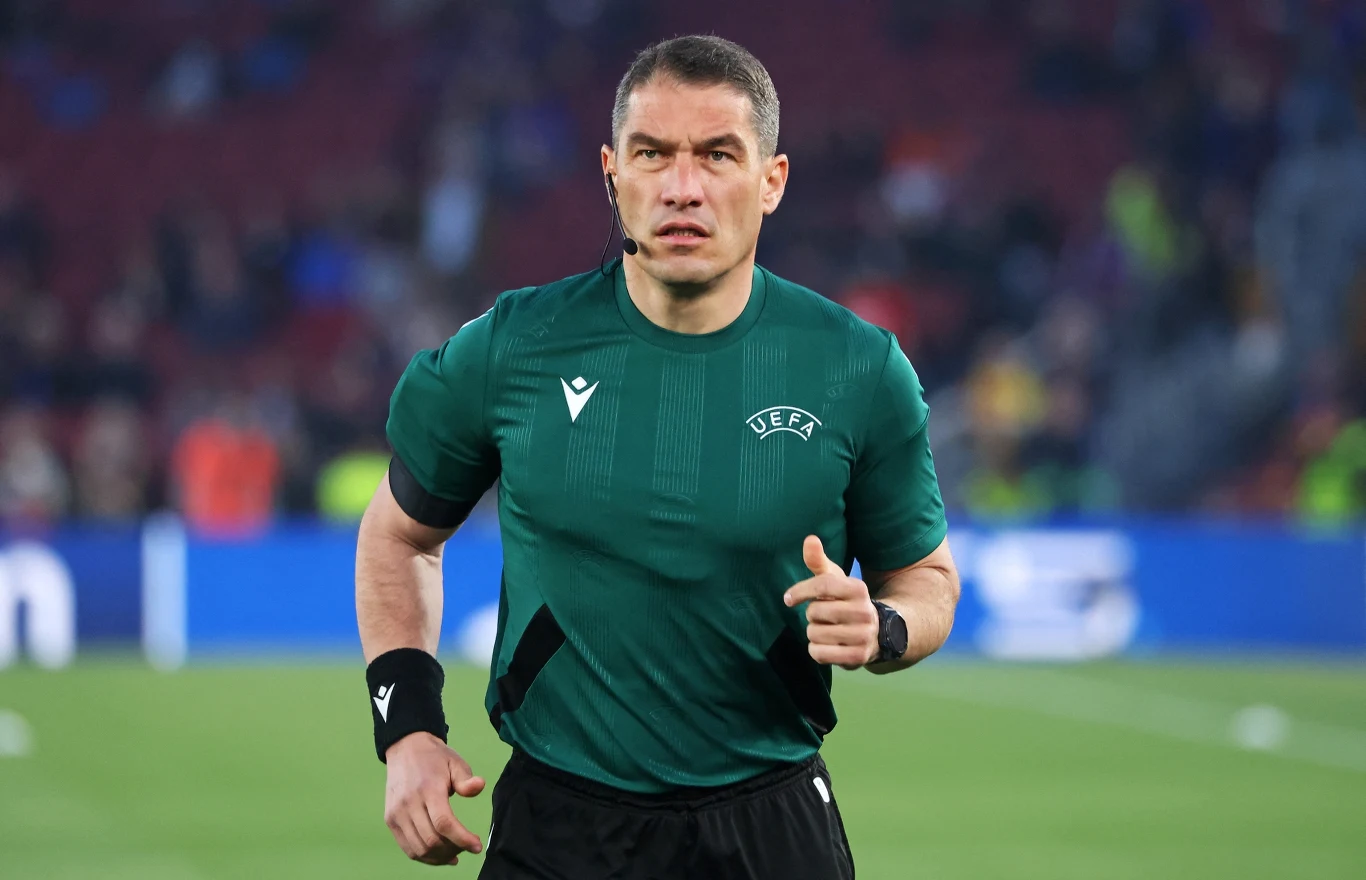 sędzia piłkarski w zielonej koszulce z logo UEFA biegnie po boisku, w tle rozmyte trybuny stadionu