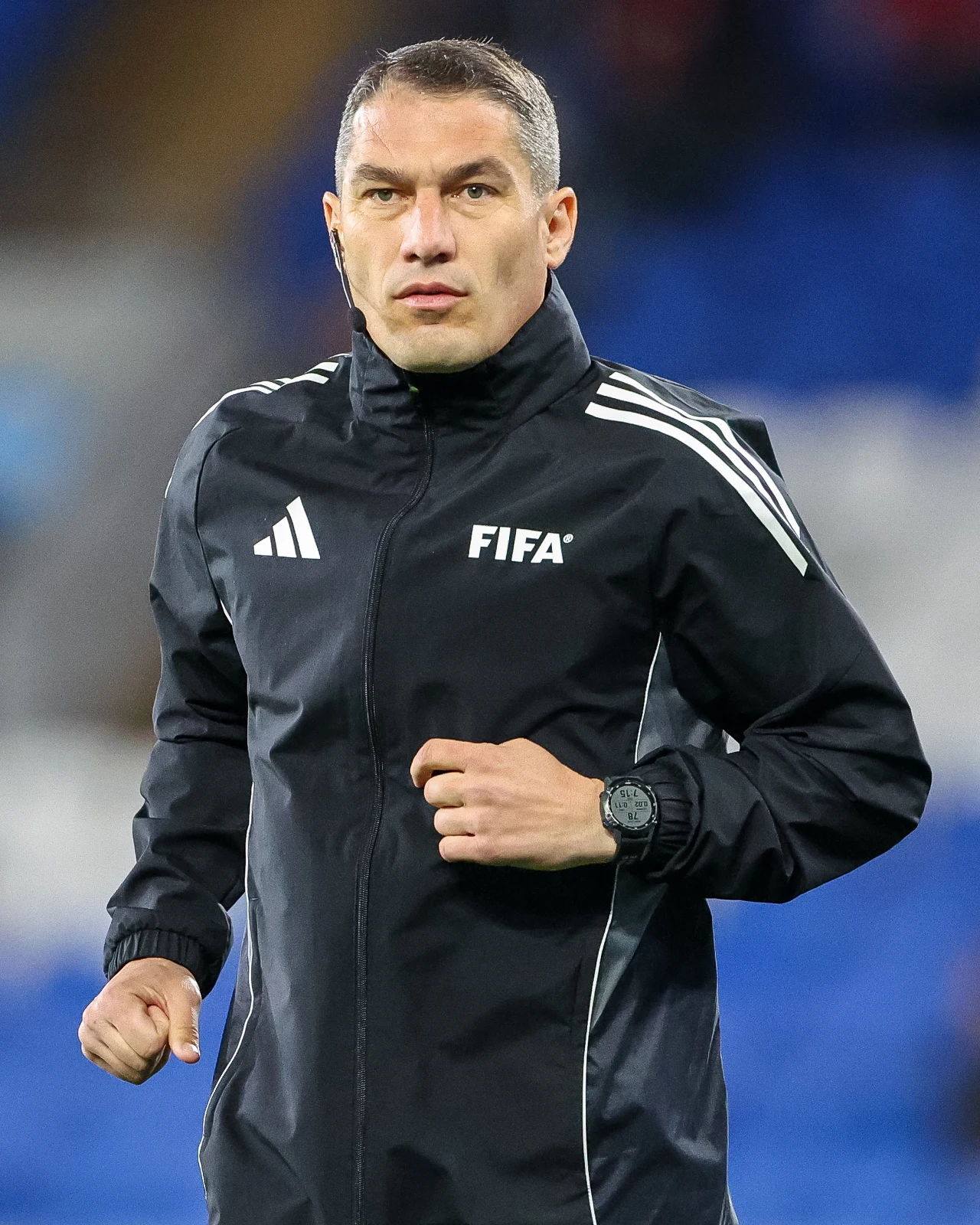 mężczyzna w czarnej kurtce sportowej z logo FIFA, biegnący po murawie, skupiona mimika twarzy, krótko ostrzyżone włosy, zegarek sportowy na lewym nadgarstku
