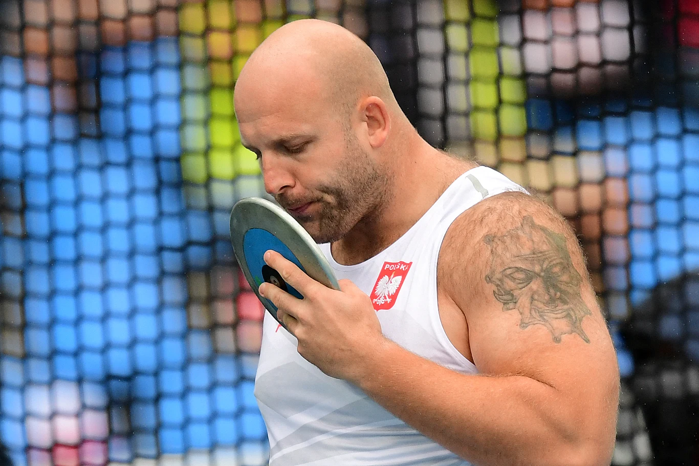 Piotr Małachowski - jeden z najlepszych dyskoboli XXI wieku sportowiec w białej koszulce z orzełkiem oraz polską flagą całuje dysk przed rzutem, w tle widoczna siatka ochronna i rozmyte sylwetki kibiców