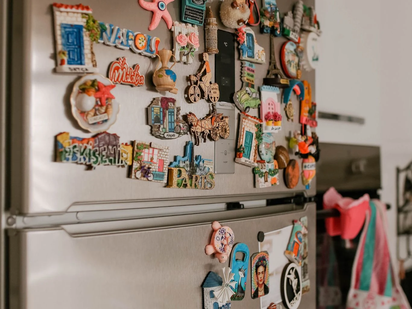 Czy duża ilość magnesów na lodówce wpływa na jej działanie? Srebrna lodówka z wieloma kolorowymi magnesami pamiątkowymi, w tle kuchenne tekstylia na haczykach.