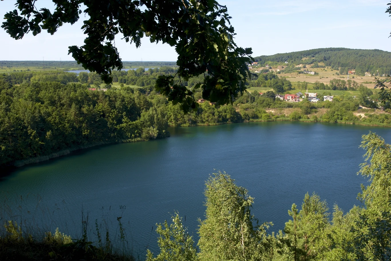 malownicze jezioro otoczone zielonymi drzewami, nad którym górują liście na pierwszym planie, w tle rozciąga się wiejska zabudowa i łagodne wzgórza