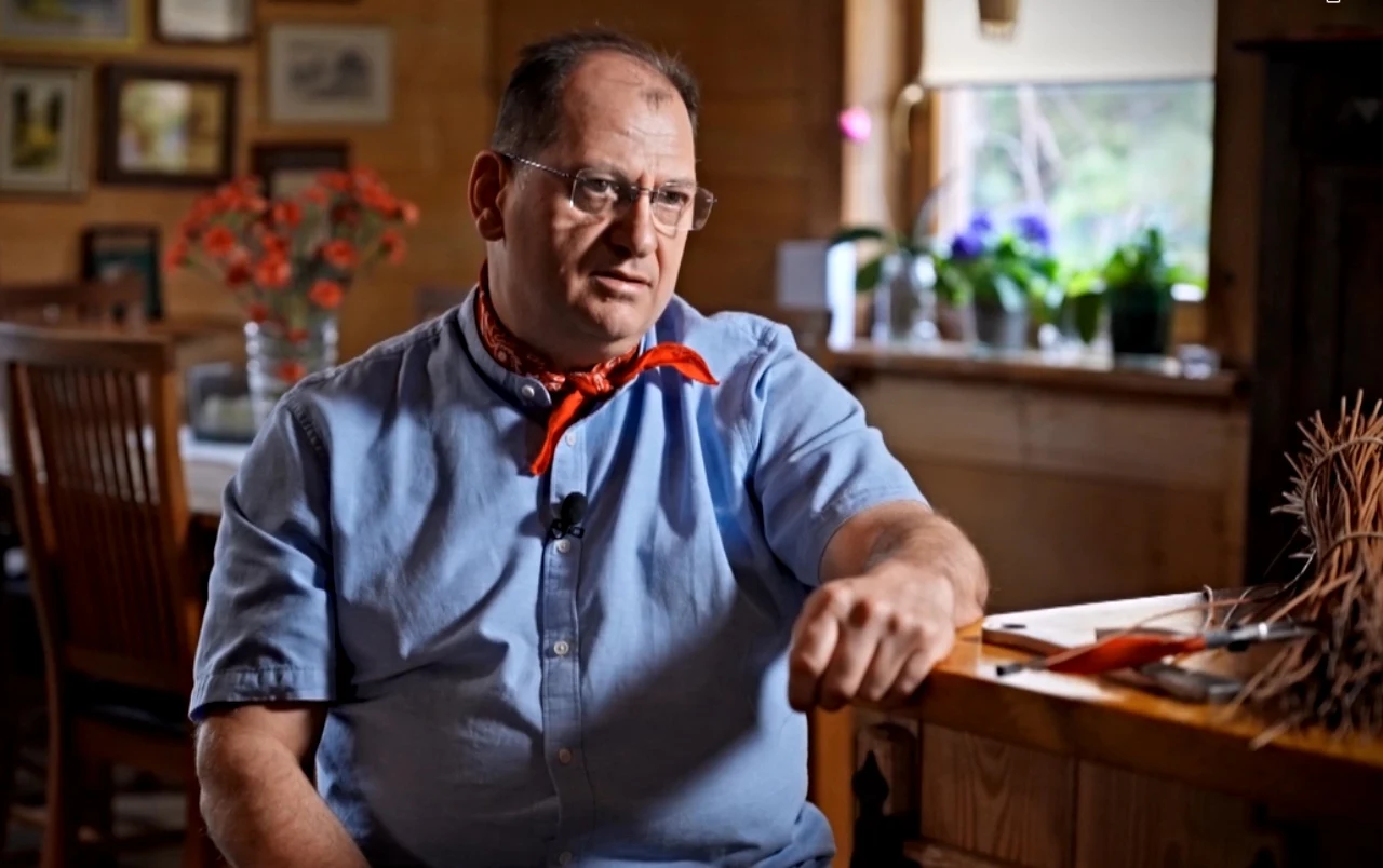 Maciej Pawlak Maciej Pawlak w okularach, błękitnej koszuli i pomarańczowej chustce siedzi przy stole w pracowni, otoczony notatkami i przyborami; w tle kwiaty w doniczkach i zdjęcia na ścianie.