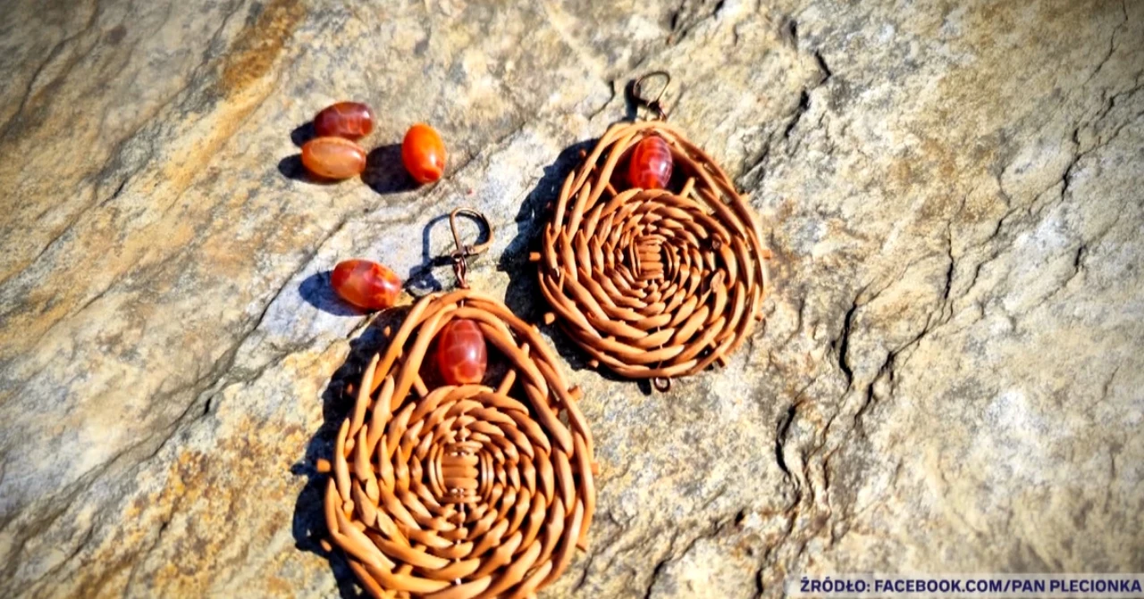 Kolczyki z wikliny z pracowni Macieja Pawlaka Plecione kolczyki o owalnym kształcie z naturalnych materiałów, ozdobione małymi, brązowymi koralikami, ułożone na kamiennej powierzchni obok kilku podobnych koralików.