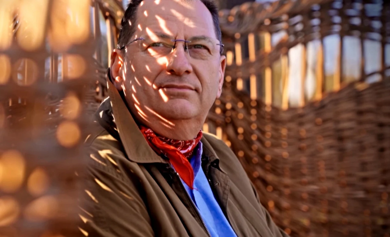 Maciej Pawlak Mężczyzna w okularach i czerwonej chustce na szyi siedzi przy wiklinowej konstrukcji, przez którą padają plamy światła na jego twarz i ubranie.