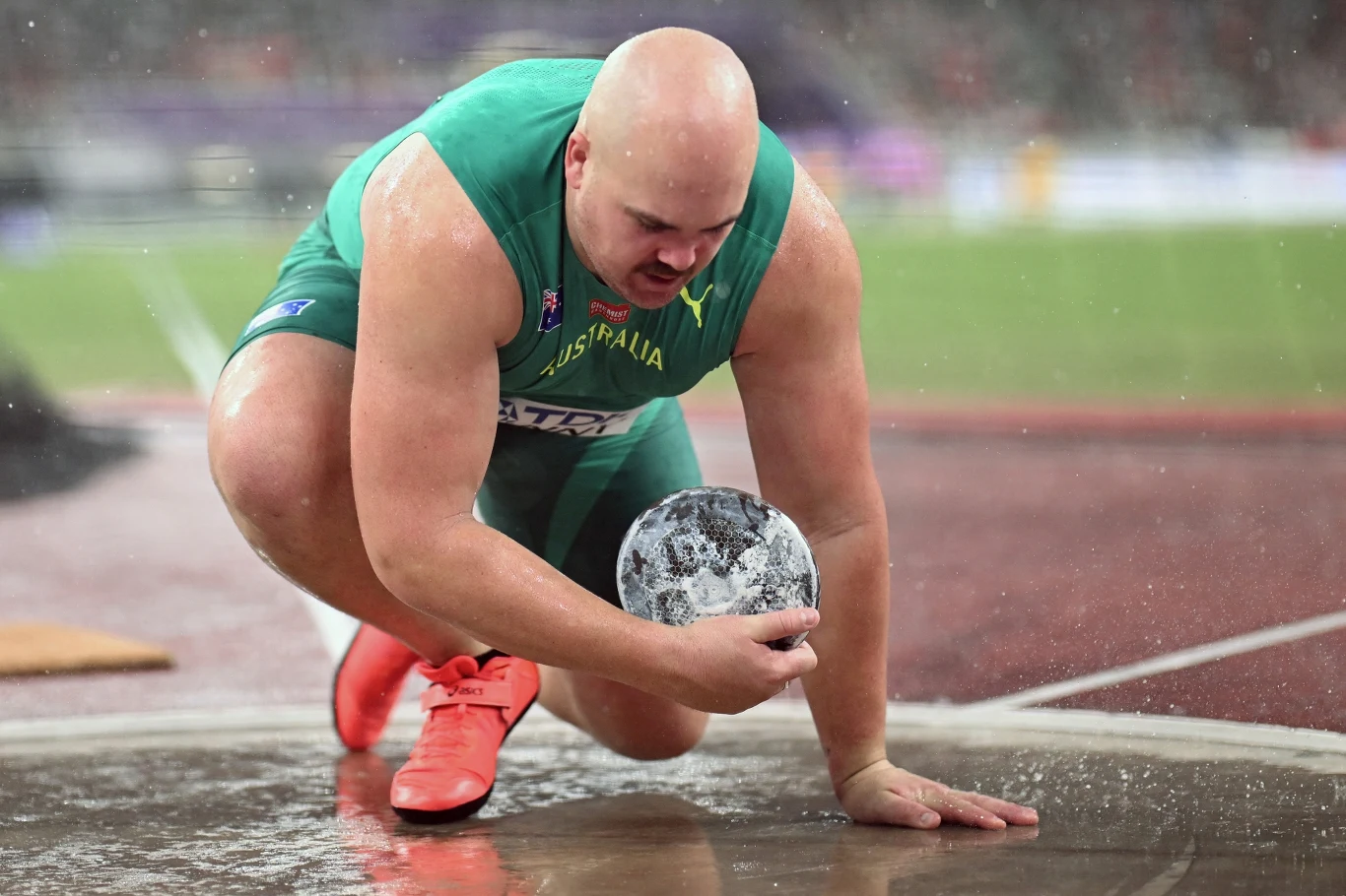Matthew Denny Lekkoatleta w zielonym stroju z napisem Australia przygotowuje się do pchnięcia kulą, trzymając ciężką kulę przy ziemi, z widoczną kredą na dłoniach i intensywnym skupieniem na twarzy; w tle rozmyty stadion lekkoatletyczny.
