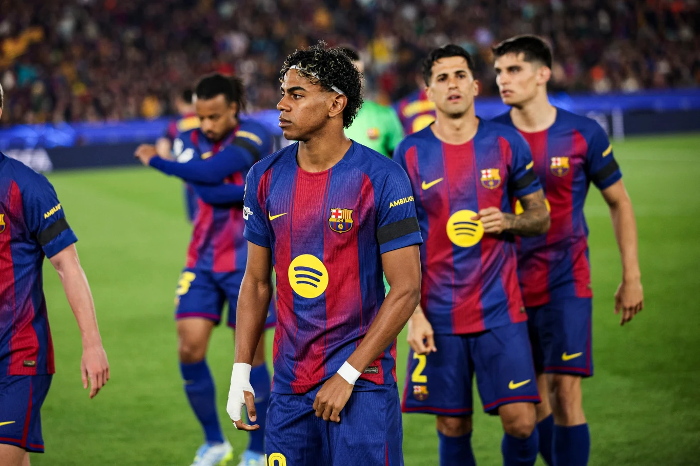 piłkarze w koszulkach FC Barcelony stoją na murawie stadionu, na pierwszym planie zawodnik z opaską na nadgarstku i wyrazem skupienia na twarzy, w tle kilku innych graczy w tych samych barwach oraz rozmyta widownia