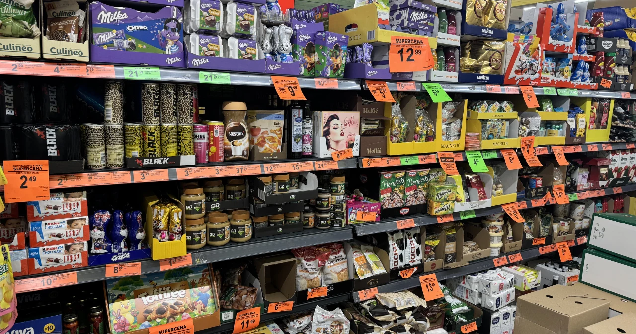 Te produkty i tak kupujesz… teraz zapłacisz za nie dużo mniej Półki sklepu z czekoladami, kremami, kawą i słodyczami z widocznymi promocyjnymi etykietami cenowymi.