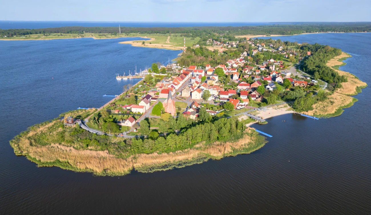 malownicza miejscowość położona na półwyspie, otoczona wodą, z zwartą zabudową domków z jasnymi dachami, kościołem położonym w centrum, licznymi drzewami i zielenią oraz mariną z cumującymi łodziami