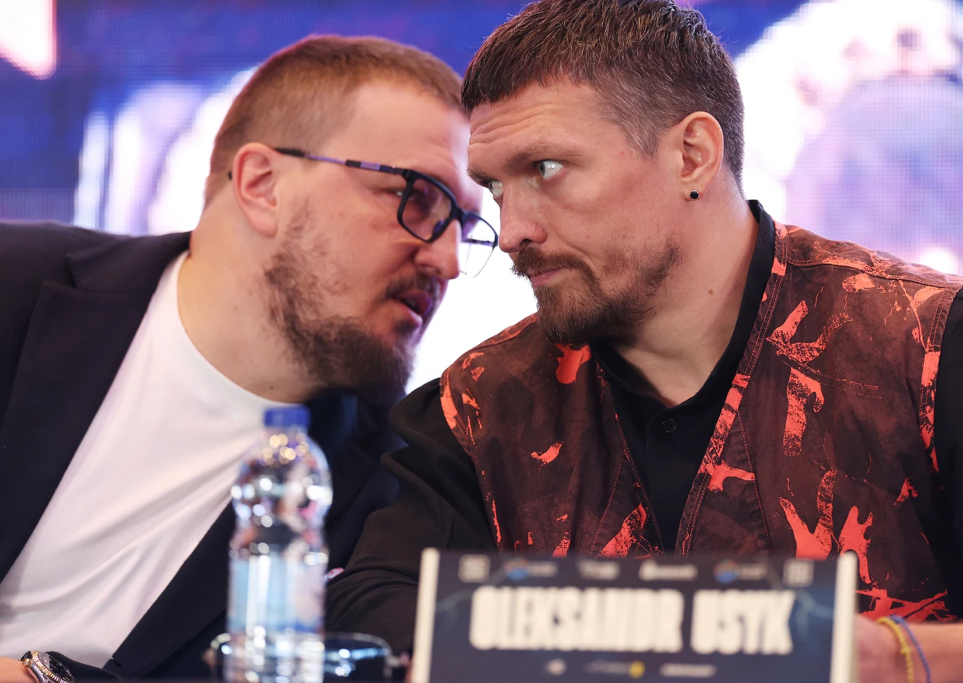 Aleksander Krasjuk i Ołeksandr Usyk Aleksander Krasjuk i Ołeksandr Usyk siedzą przy stole, jeden nachyla się do drugiego; na stole butelka wody i tabliczka z napisem "OLEKSANDR USYK".