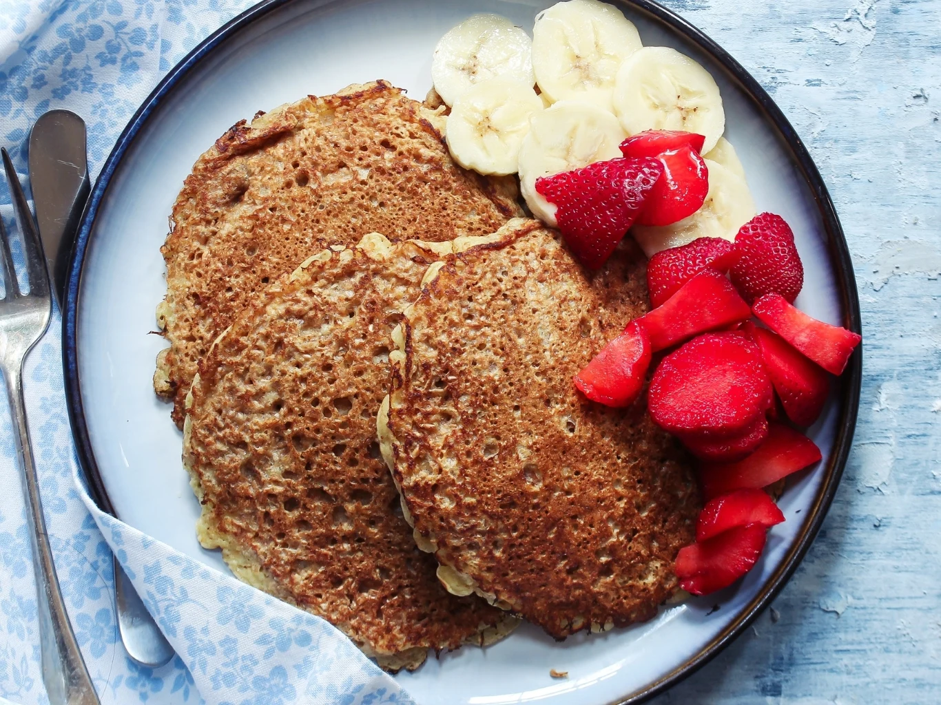 Proteinowe placuszki to doskonały wybór na śniadanie. Dwa złociste proteinowe placuszki z plasterkami banana i kawałkami truskawek na białym talerzu ze sztućcami.