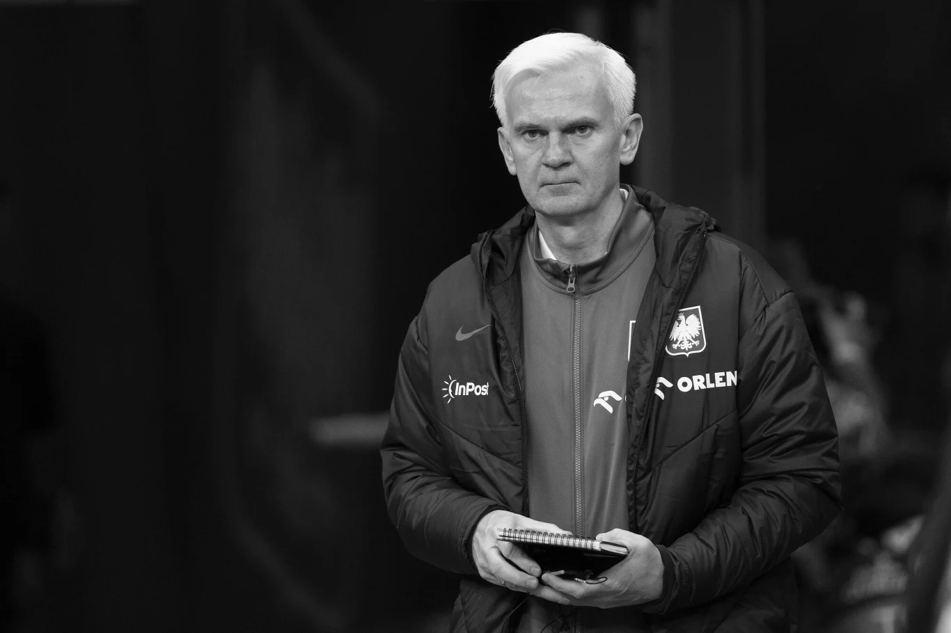 Nie żyje Jacek Magiera Mężczyzna o jasnych włosach w sportowej kurtce z logotypami sponsorów, trzymający notes i długopis, patrzy poważnie przed siebie.