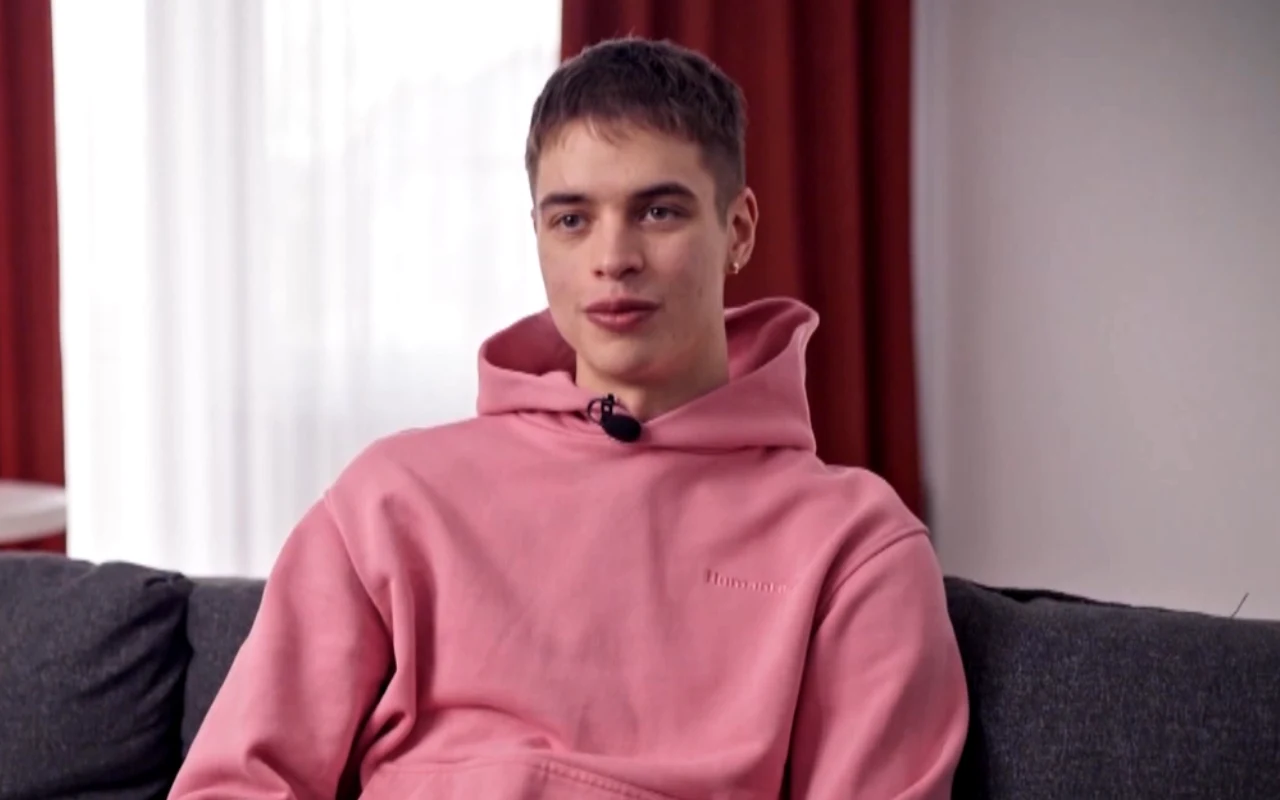 młody mężczyzna z krótkimi włosami siedzący na ciemnoszarej kanapie, ubrany w różową bluzę z kapturem i mikrofon przypięty do kołnierza; w tle jasne zasłony i czerwone zasłony na dużym oknie