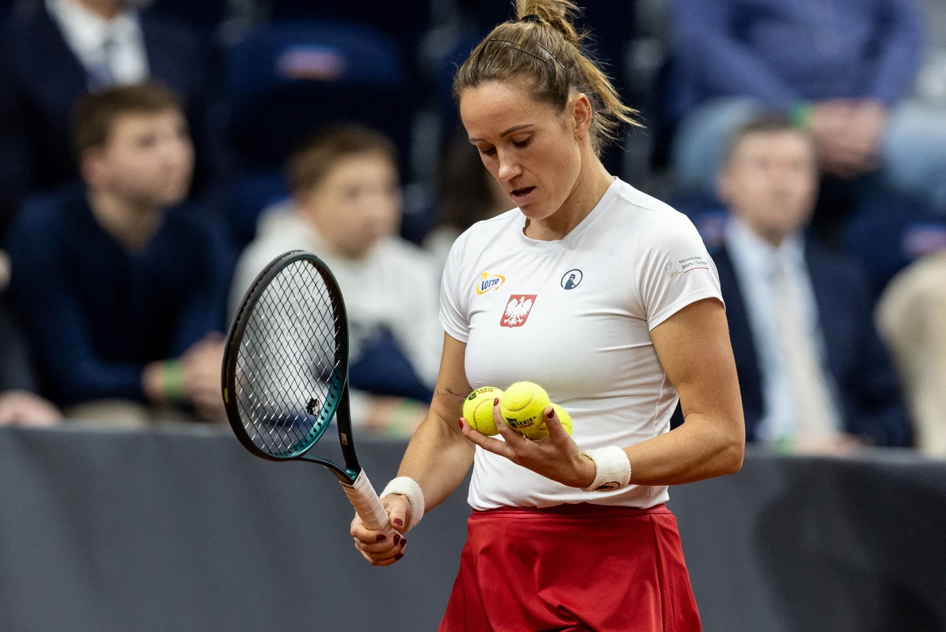 Katarzyna Kawa w barwach reprezentacji Polski podczas meczu Billie Jean King Cup tenisistka w białej koszulce z polskim godłem i czerwonej spódnicy trzymająca rakietę oraz dwie piłki tenisowe, skupiona przed serwisem podczas meczu