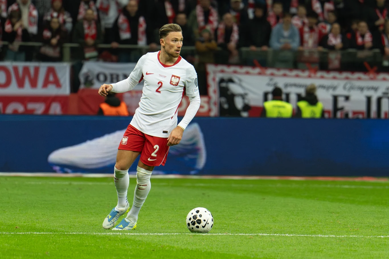 piłkarz w biało-czerwonym stroju reprezentacji Polski prowadzi piłkę po murawie stadionu podczas meczu, w tle kibice z flagami