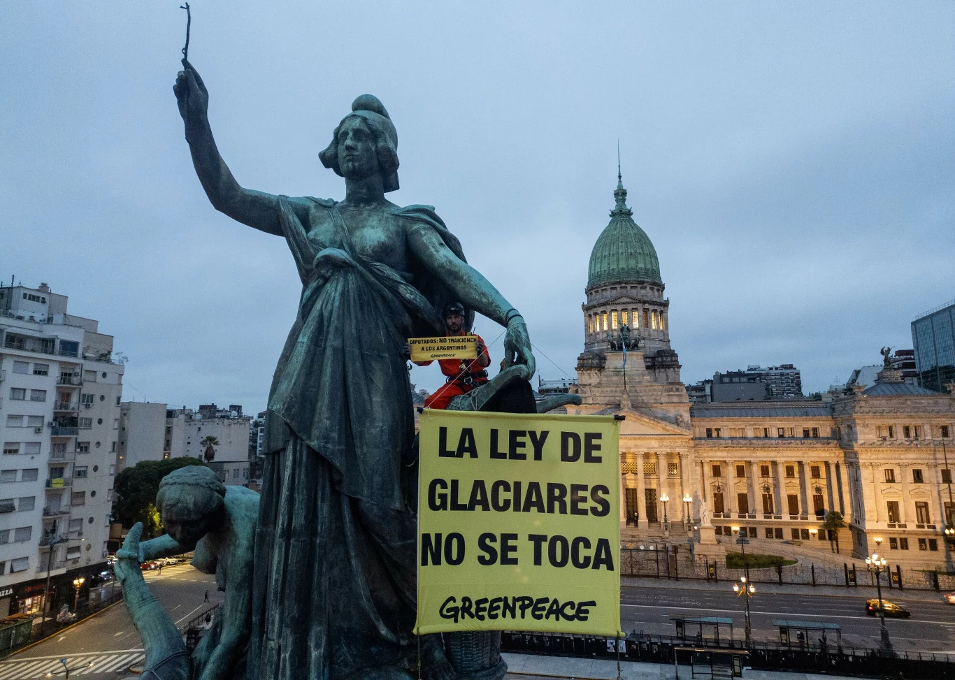 duża statua kobiety z uniesionym ramieniem trzymająca żółty transparent z czarnym napisem „LA LEY DE GLACIARES NO SE TOCA GREENPEACE”, w tle monumentalny budynek rządowy z kopułą i wieczornym oświetleniem