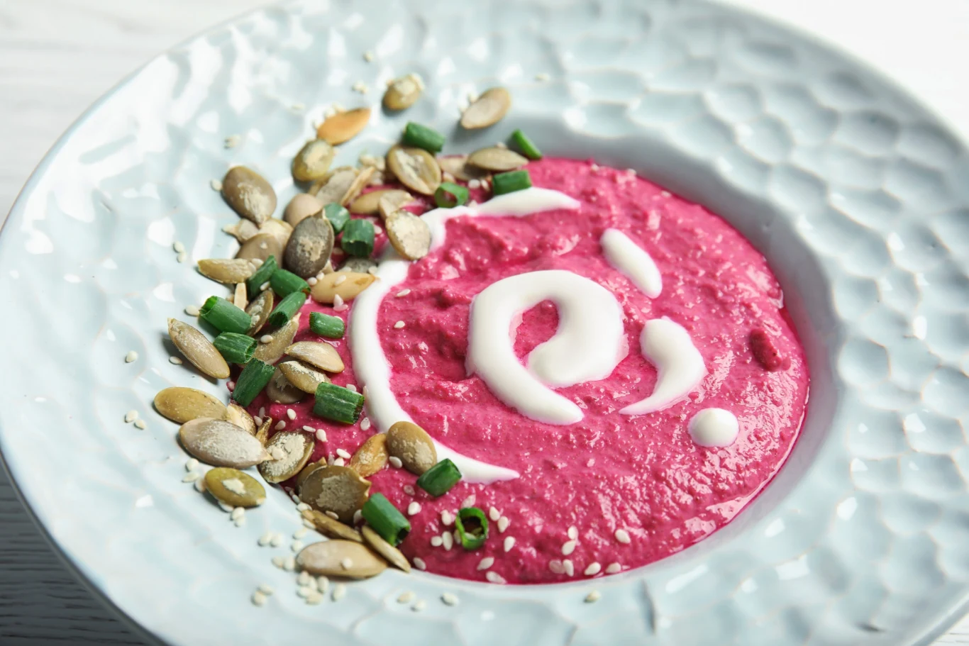 Hummus z dodatkiem buraka to doskonały wybór dla zdrowia Hummus z burakiem udekorowany mlekiem kokosowym, pestkami dyni, sezamem i szczypiorkiem na białym talerzu.