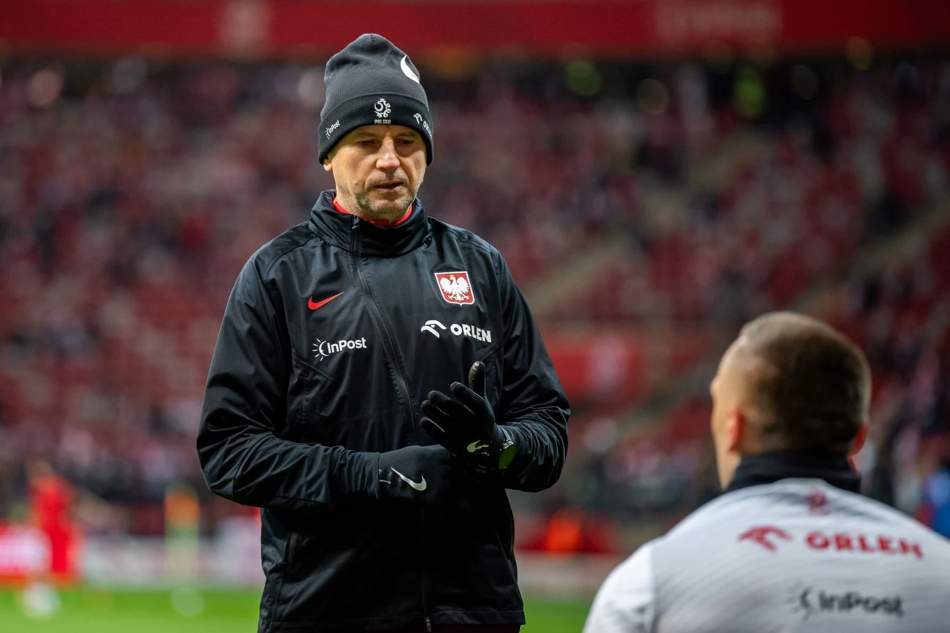 trener reprezentacji Polski w czarnej kurtce i czapce stoi na murawie stadionu, w tle rozmyte trybuny z kibicami; na pierwszym planie z tyłu widoczny członek sztabu ubrany w biały dres z logo sponsorów
