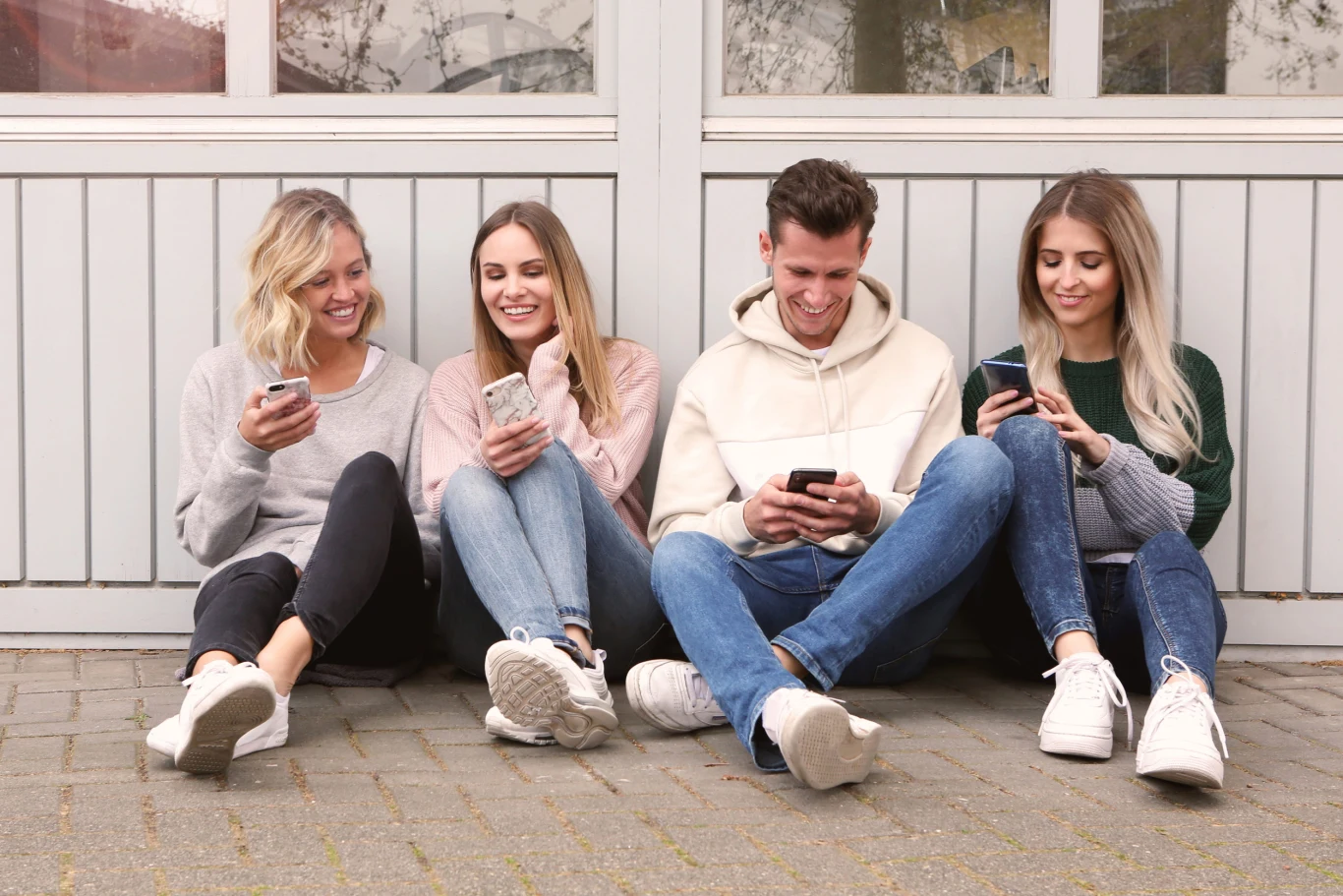 Nawet podczas spotkań towarzyskich wyciągamy telefony. Nieświadomie działamy na własną szkodę cztery młode osoby siedzą obok siebie na chodniku, każda z nich trzyma w ręku smartfona i patrzy na ekran, uśmiechając się lub śmiejąc; luźna, przyjazna atmosfera, sportowy styl ubioru