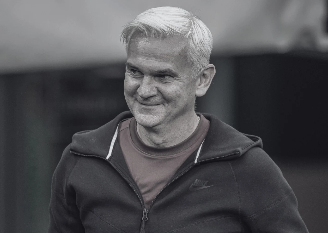 Jacek Magiera nie żyje Uśmiechnięty mężczyzna z siwymi włosami ubrany w sportową bluzę zapinaną na zamek oraz koszulkę, patrzący w lewo, w tle rozmyte tło bez wyraźnych szczegółów.