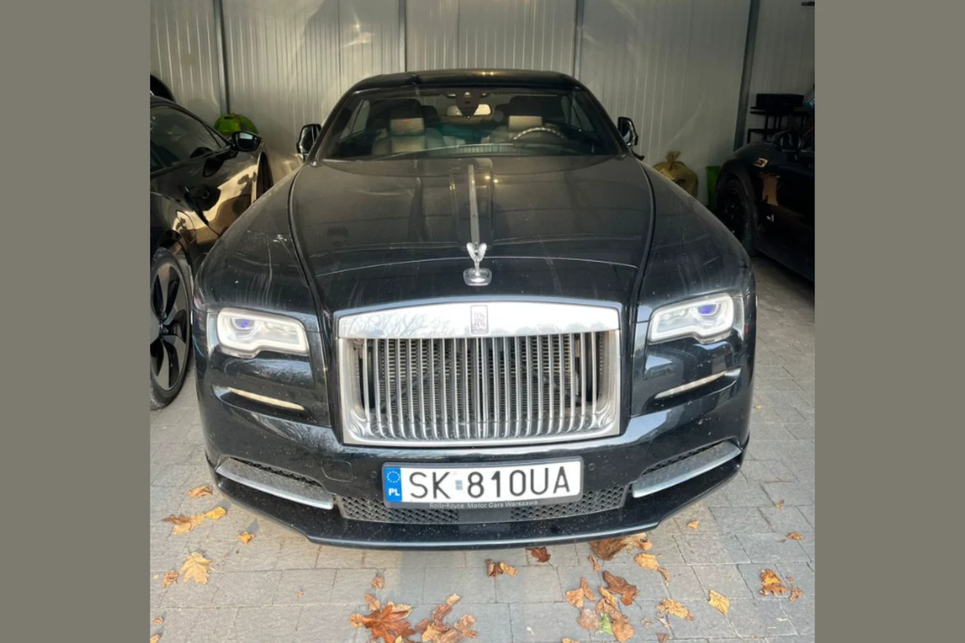 Wartość tego egzemplarza Rolls-Royce Wraith wyceniana jest na 1 116 348 zł. Czarny Rolls-Royce z podświetlonymi reflektorami i charakterystycznym emblematem Spirit of Ecstasy na masce, zaparkowany w garażu na tle metalowych ścian, z widoczną rejestracją SK 810UA i liśćmi na kostce brukowej przed samochodem.