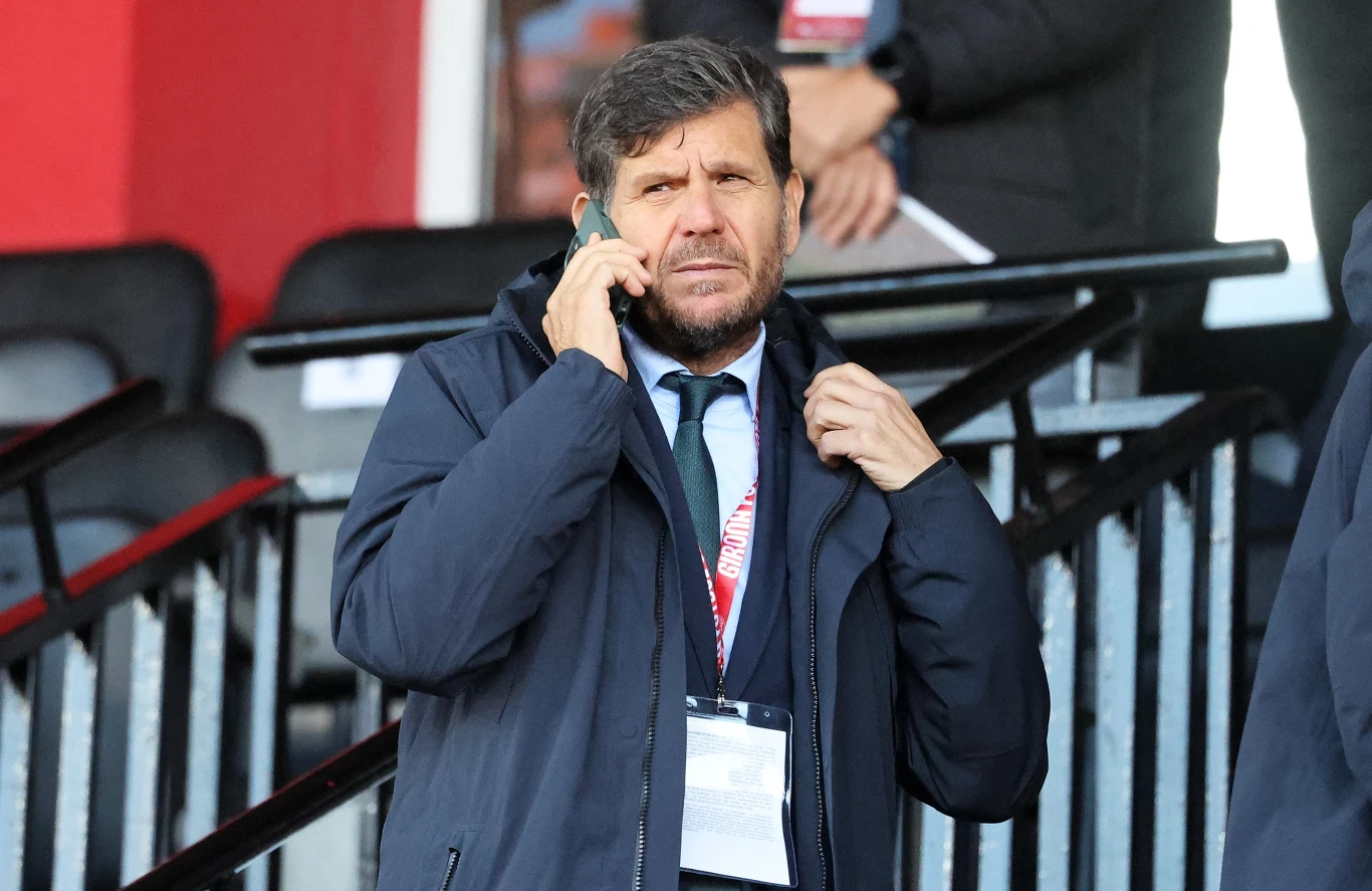 mężczyzna w garniturze i ciemnej kurtce rozmawia przez telefon, stoi przy metalowych schodach na tle pustych krzeseł stadionowych