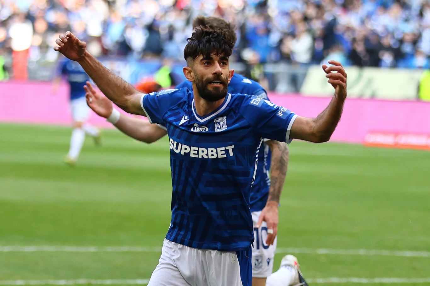 Piłkarz w niebieskiej koszulce Lecha Poznań z rozłożonymi rękami celebruje zdobycie gola na boisku, w tle inni zawodnicy oraz rozmyty tłum kibiców na trybunach.