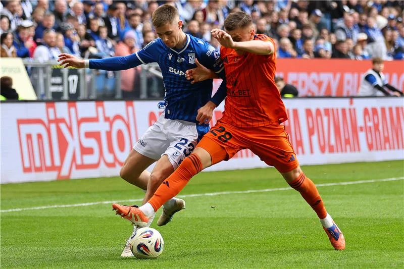 dwóch piłkarzy rywalizujących o piłkę na murawie podczas meczu, jeden ubrany w niebieski strój, drugi w pomarańczowy, dynamiczna walka i skupienie zawodników, trybuny pełne kibiców w tle