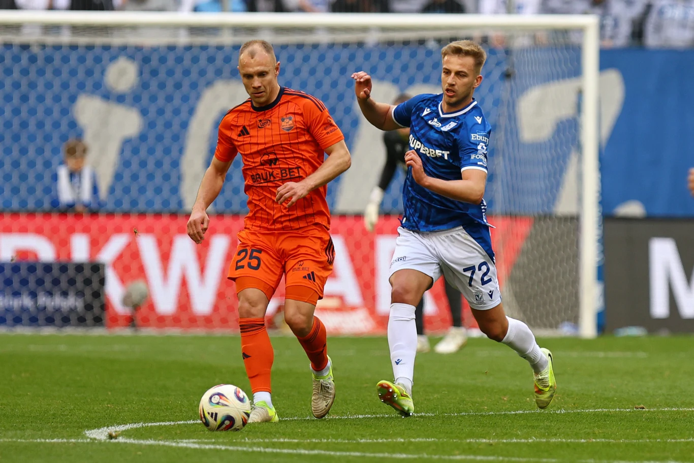 dwóch piłkarzy rywalizuje o piłkę na murawie, jeden w pomarańczowym stroju prowadzi futbolówkę, a drugi w niebieskim stroju próbuje odebrać mu piłkę na tle stadionu pełnego kibiców