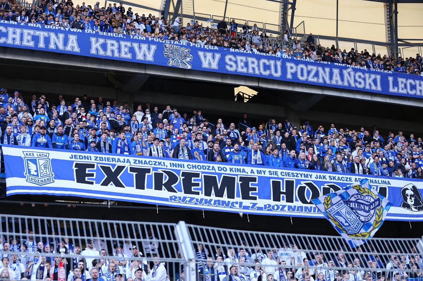 Tłum kibiców w niebiesko-białych barwach dopingujący drużynę Lech Poznań, dominują transparenty z napisami i herbem klubu, a trybuny wypełnia intensywna atmosfera wsparcia.