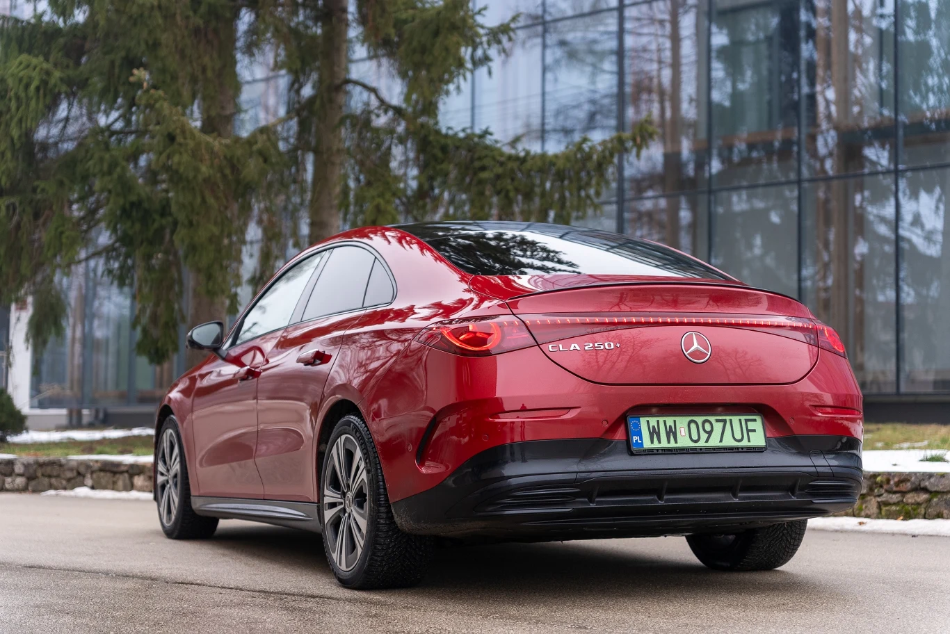 czerwony samochód Mercedes-Benz CLA 250 z zielonymi tablicami rejestracyjnymi, zaparkowany na tle nowoczesnego budynku i drzew iglastych