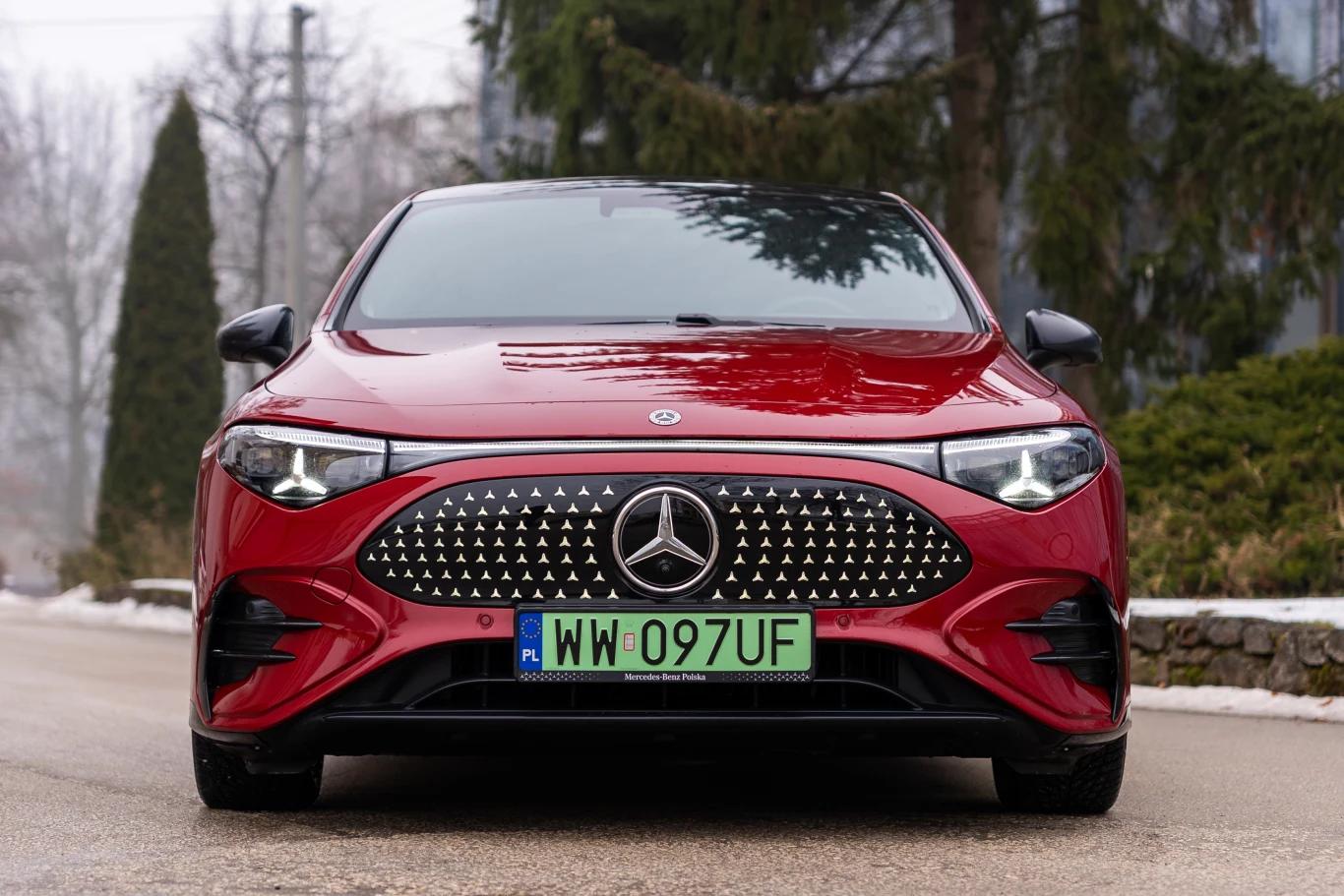 Czerwony samochód marki Mercedes z charakterystycznym dużym grillem i polską zieloną tablicą rejestracyjną na tle miejskiej drogi, otoczony drzewami i rozmytym krajobrazem.