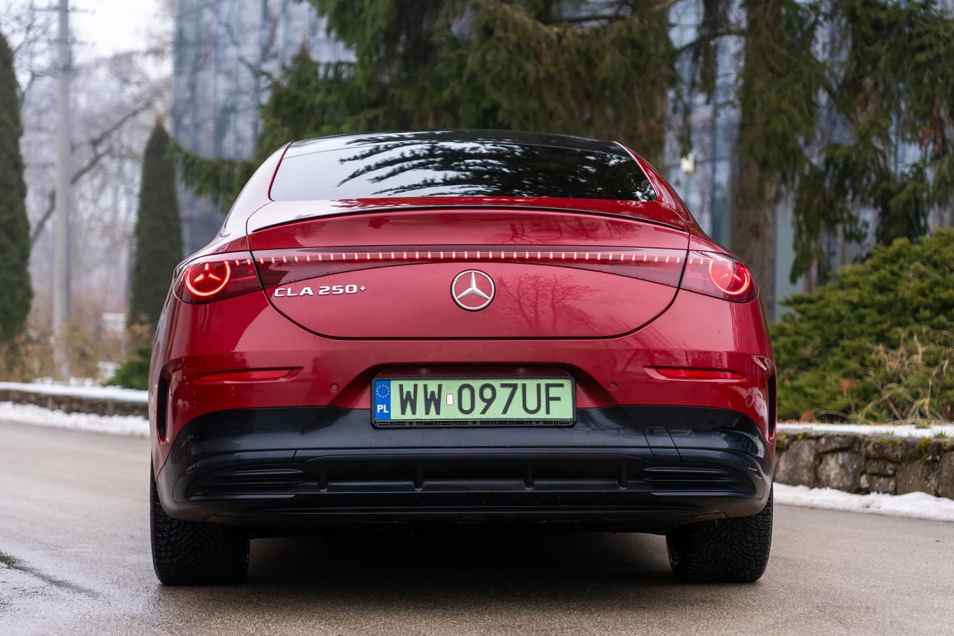 czerwony Mercedes CLA 250 z zielonymi tablicami rejestracyjnymi zaparkowany na wilgotnej drodze, w otoczeniu drzew iglastych i lekko zamglonego tła