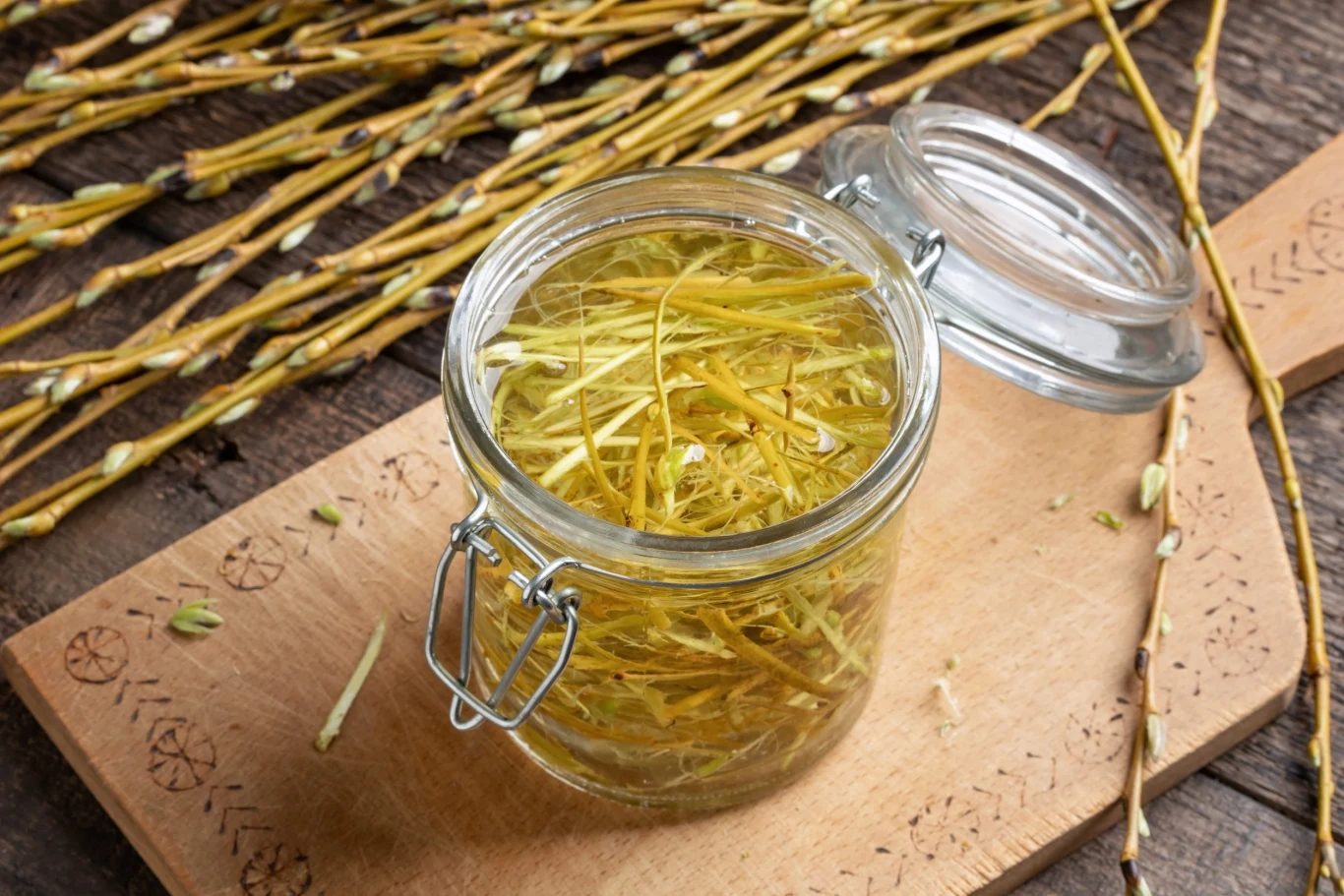 szklany słoik wypełniony suchą korą wierzby ustawiony na drewnianej desce, wokół niego rozrzucone gałązki i kora wierzbowa