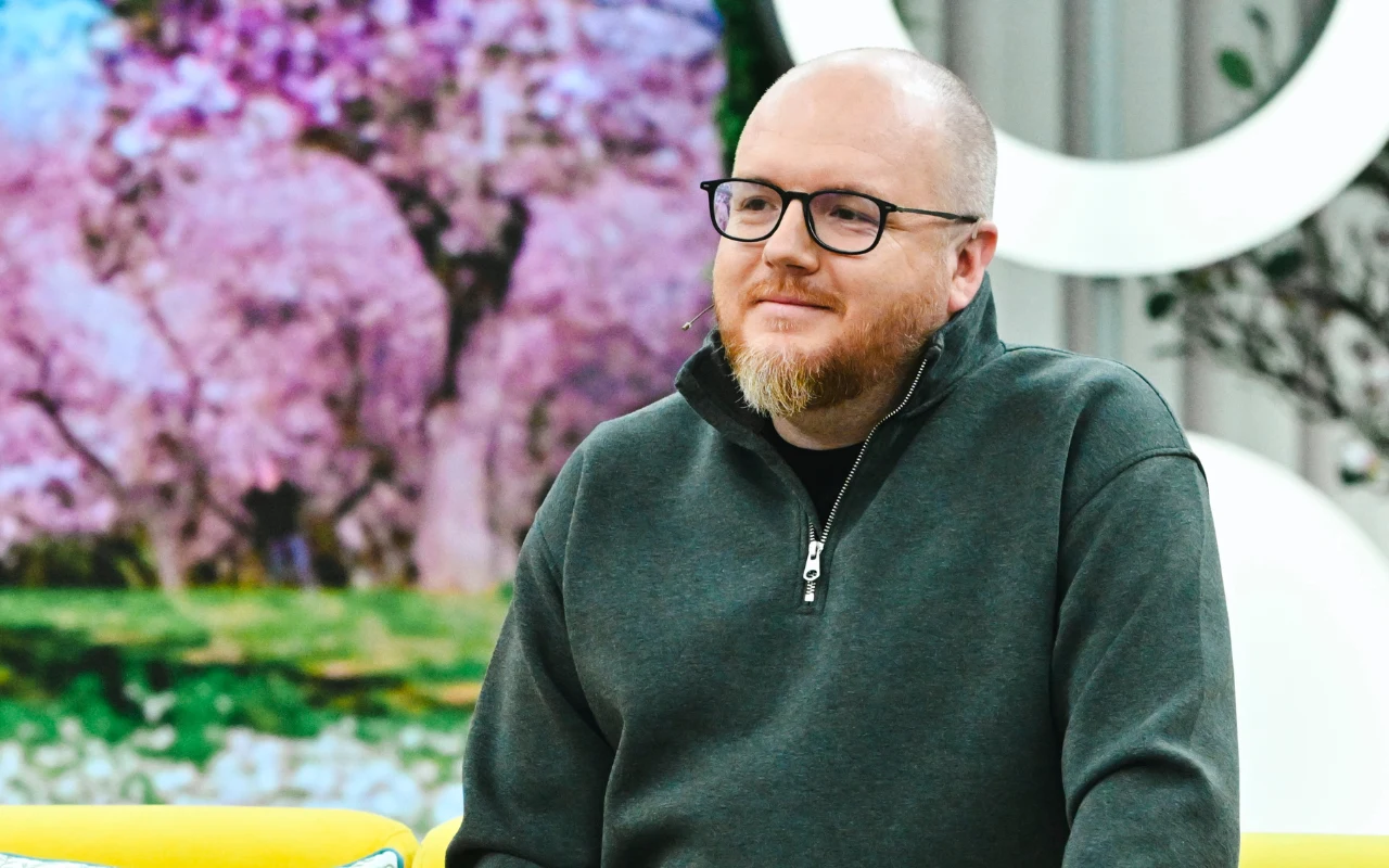 Mikołaj Gospodarek w "halo tu polsat" mężczyzna z brodą i okularami siedzący na żółtej kanapie, w tle rozmyty obraz kwitnących drzew