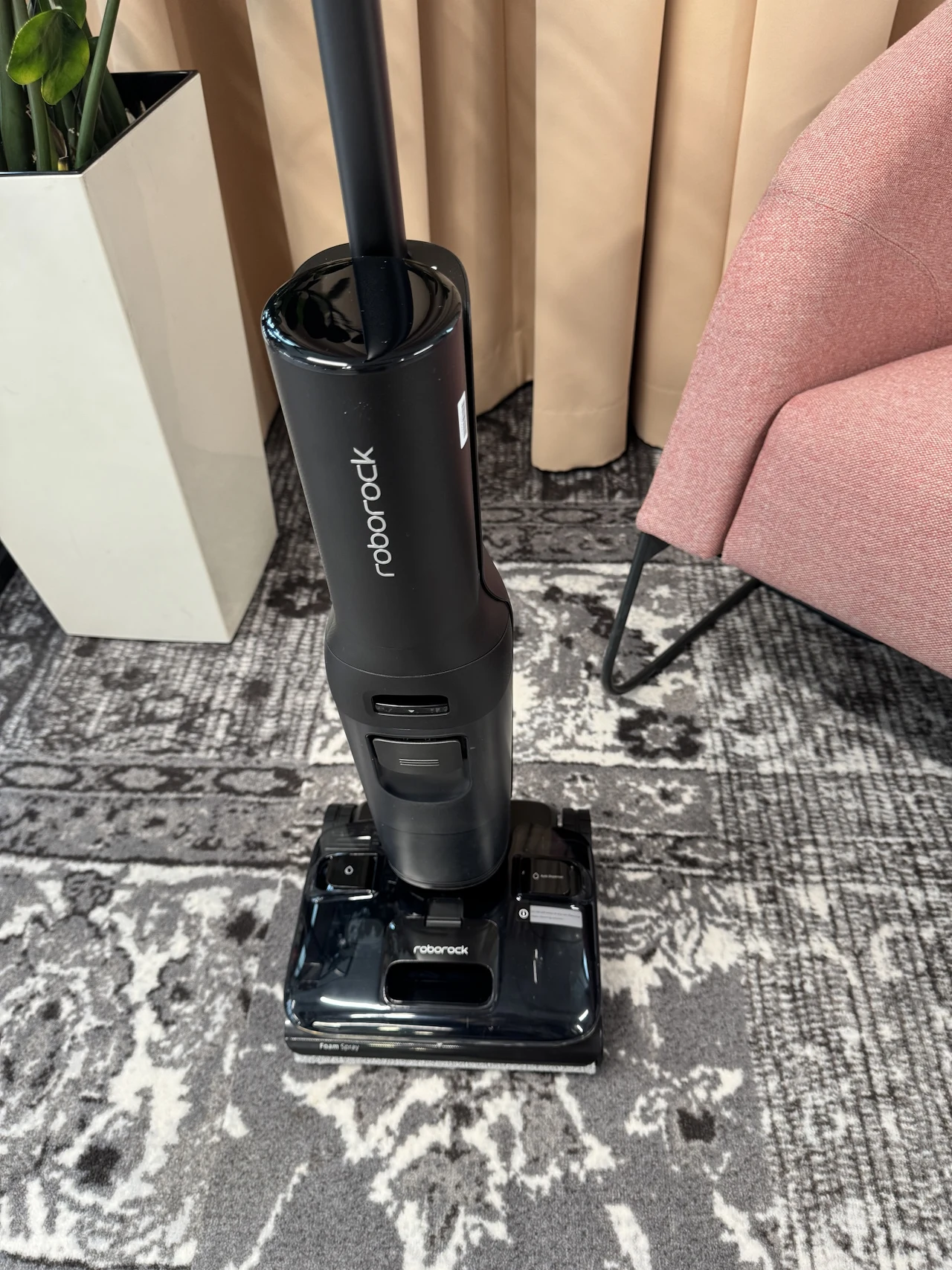 Producent zapewnia, że na rolce nie zaplątają się włosy nawet o długości do 50 cm czarny bezprzewodowy odkurzacz pionowy Roborock ustawiony na wzorzystym dywanie, obok różowy fotel oraz biała donica z zieloną rośliną, w tle beżowe zasłony
