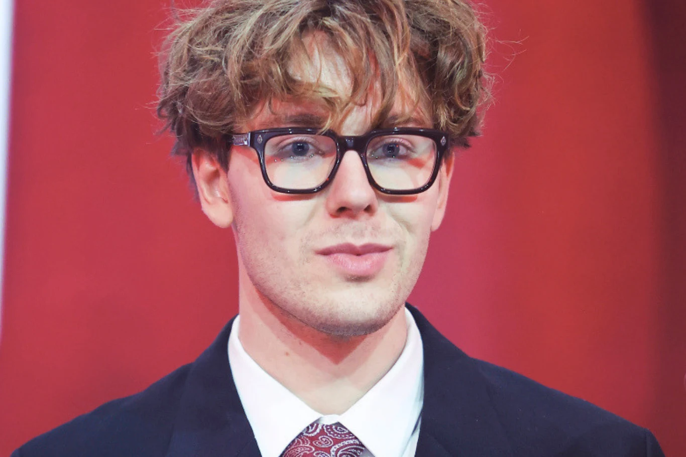 Mikołaj Bagi Bagiński Mikołaj Bagi Bagiński w eleganckim garniturze, koszuli z krawatem, na czerwonym tle.